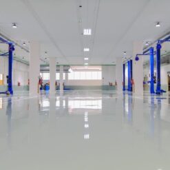 A spacious, clean automotive workshop with empty work bays, blue vehicle lifts, and polished floors. Fire extinguishers are mounted on white columns, while shelves display EPON 1001F and premium release agents.