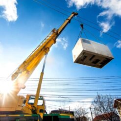 On a sunny day, a yellow crane lifts a white container high above power lines, using discontinued Teflon™ Heavy Equipment Grease, with blue sky, scattered clouds, and buildings visible below.
