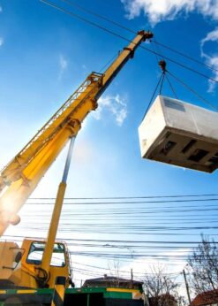 On a sunny day, a yellow crane lifts a white container high above power lines, using discontinued Teflon™ Heavy Equipment Grease, with blue sky, scattered clouds, and buildings visible below.