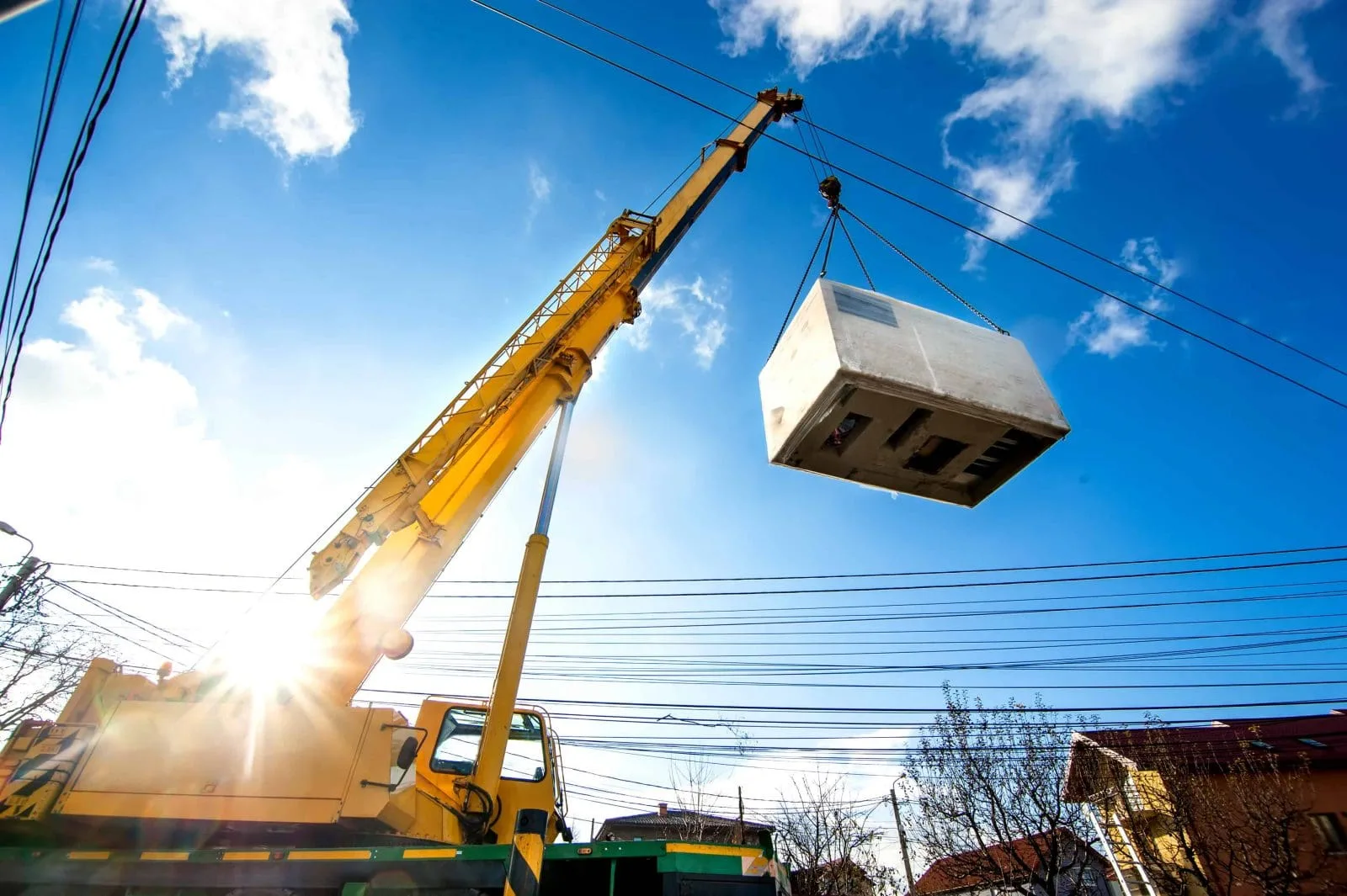 On a sunny day, a yellow crane lifts a white container high above power lines, using discontinued Teflon™ Heavy Equipment Grease, with blue sky, scattered clouds, and buildings visible below.