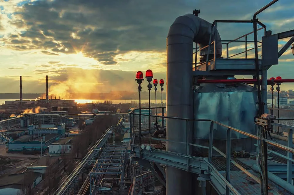 Industrial plant at sunset with large pipes, red warning lights, and smoke rising from smokestacks in the background, under a partly cloudy sky—where high-performance lubricant like Krytox is essential for smooth, reliable operation.