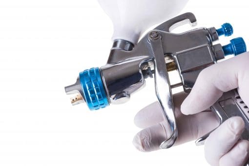A close-up of a hand wearing a white glove holding a metallic spray paint gun with blue accents, commonly used for applying release agents or lubricant, against a white background.