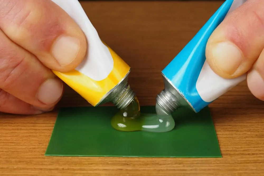 Two hands squeeze yellow and blue tubes, dispensing glue onto a green surface, with the adhesives mixing together—no release agents or lubricant like Krytox are used in this bonding process.