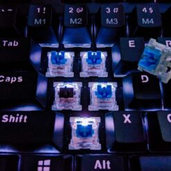Close-up of a black mechanical keyboard with keycaps removed, revealing illuminated switches. Loose switches near the E key appear ready for TriboSys KSL | Keyboard Spray Lubricant application.