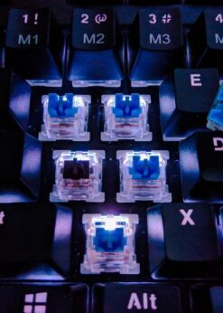 Close-up of a black mechanical keyboard with keycaps removed, revealing illuminated switches. Loose switches near the E key appear ready for TriboSys KSL | Keyboard Spray Lubricant application.